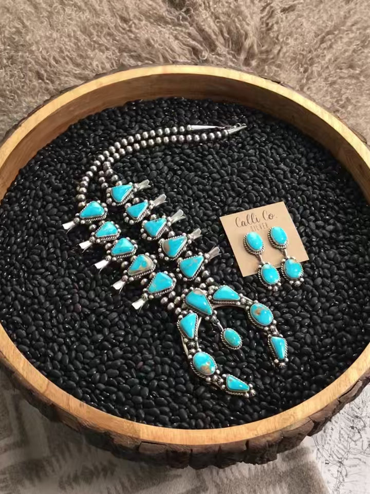 The Reign Squash Blossom Necklace Set from Calli Co Silver. Displayed on a black textured backdrop rotating on a display in a circle. Squash blossom necklace with turquoise stones and sterling silver pearl beads. Shown with coordinating dangle earrings.  